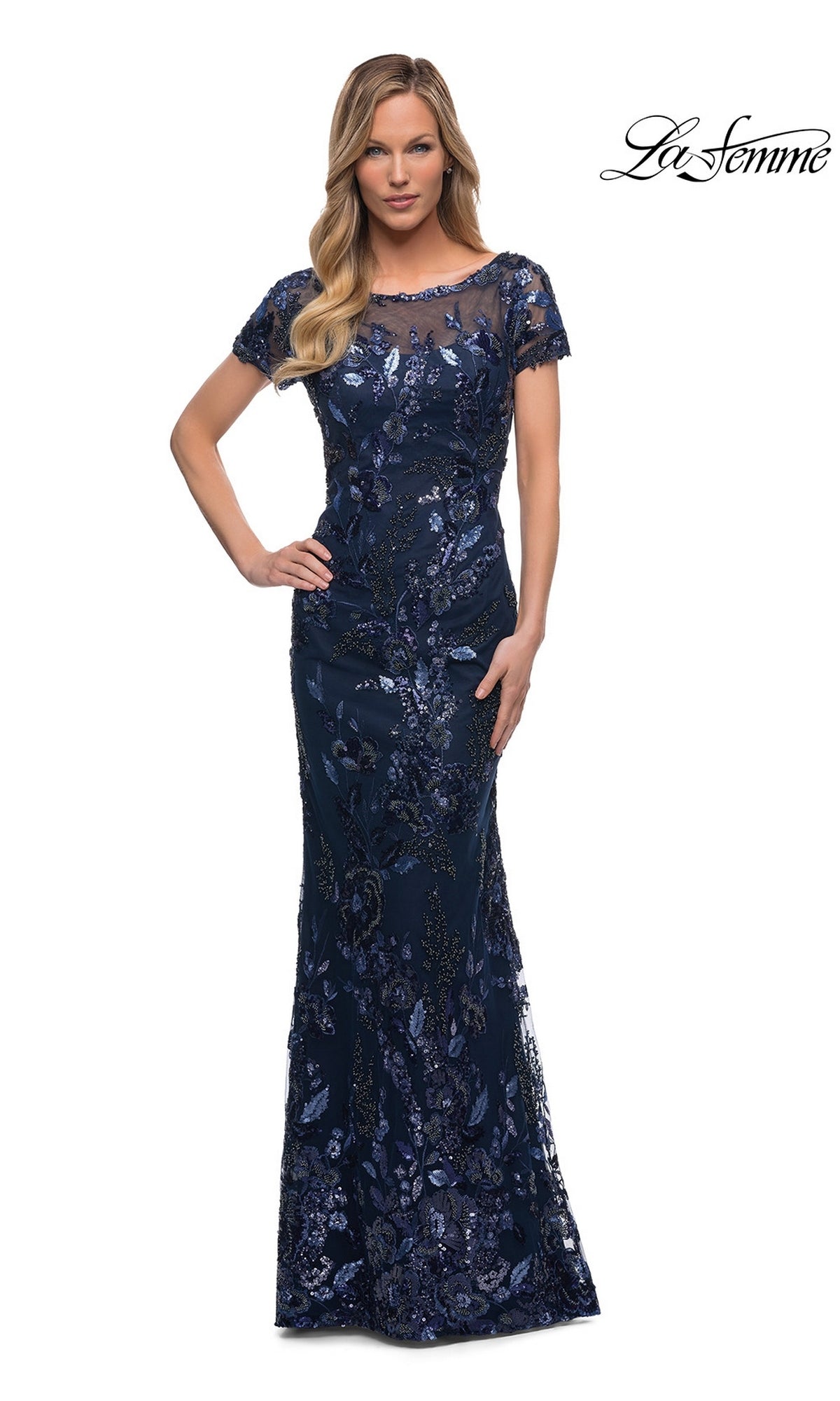 A woman in a fitted navy sheath covered in floral sequin embroidery poses with one hand on her hip. Long Formal Dress: La Femme 29961.